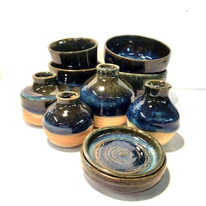 Könnte beinhalten: Eine Sammlung handgefertigter Keramikwaren mit glänzender, dunkelblauer Glasur. Das Set umfasst kleine Vasen, Schalen und Teller mit einem natürlichen, hellbraunen Sockel. Die Keramik hat eine rustikale, handwerkliche Ästhetik.