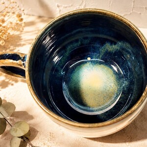 May include: A ceramic mug with a wide, circular opening and a sturdy handle. The interior features a deep navy blue glaze that transitions into a bright, speckled yellow centre. The rim and handle have a rustic, earthy brown finish.