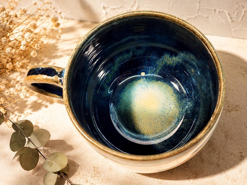 May include: A ceramic mug with a wide, circular opening and a sturdy handle. The interior features a deep navy blue glaze that transitions into a bright, speckled yellow centre. The rim and handle have a rustic, earthy brown finish.