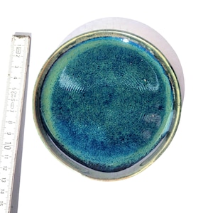 May include: A handmade ceramic dish with a teal and blue glaze. The dish has a rounded shape with a slightly raised edge. The rim is a dark bronze color. The dish is approximately 6 inches in diameter.