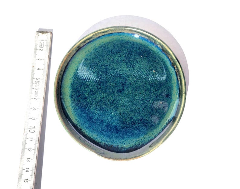 May include: A handmade ceramic dish with a teal and blue glaze. The dish has a rounded shape with a slightly raised edge. The rim is a dark bronze color. The dish is approximately 6 inches in diameter.