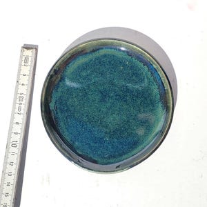 May include: A handmade ceramic plate with a glossy, textured surface. The plate features a blend of teal and blue hues, with a dark rim. The plate's diameter is approximately 6 inches.