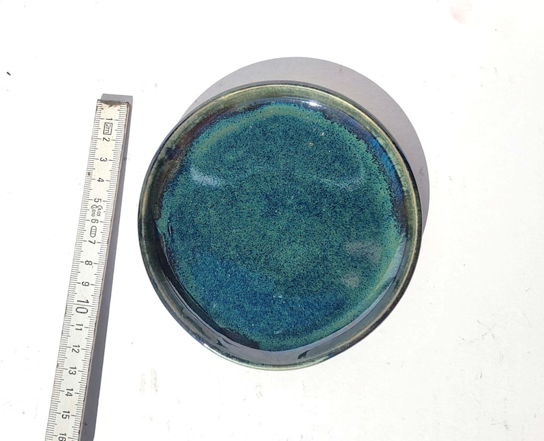 May include: A handmade ceramic plate with a glossy, textured surface. The plate features a blend of teal and blue hues, with a dark rim. The plate's diameter is approximately 6 inches.