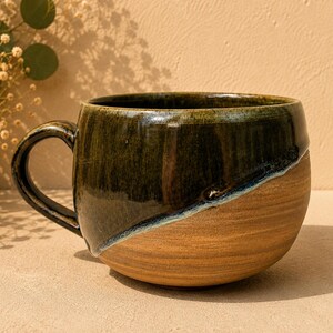 May include: A ceramic mug featuring a rounded shape and a handle. The upper portion is finished with a glossy, dark forest green glaze, while the lower section remains in a natural, unglazed tan clay finish, separated by a diagonal line.