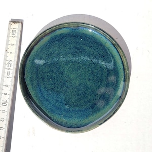 May include: A handmade ceramic plate with a textured, teal and blue glaze. The plate has a dark rim and measures approximately 6 inches in diameter. A wooden ruler is placed next to the plate for scale.