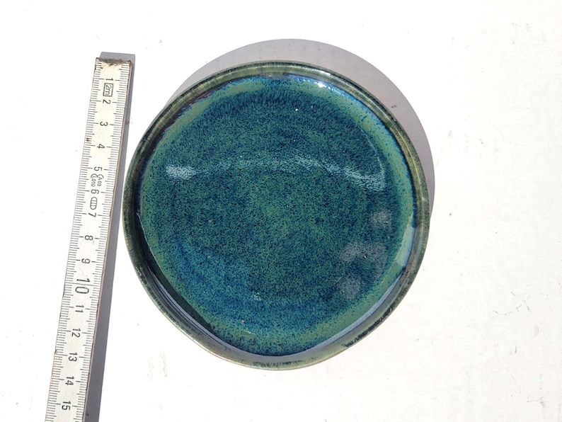 May include: A handmade ceramic plate with a textured, teal and blue glaze. The plate has a dark rim and measures approximately 6 inches in diameter. A wooden ruler is placed next to the plate for scale.