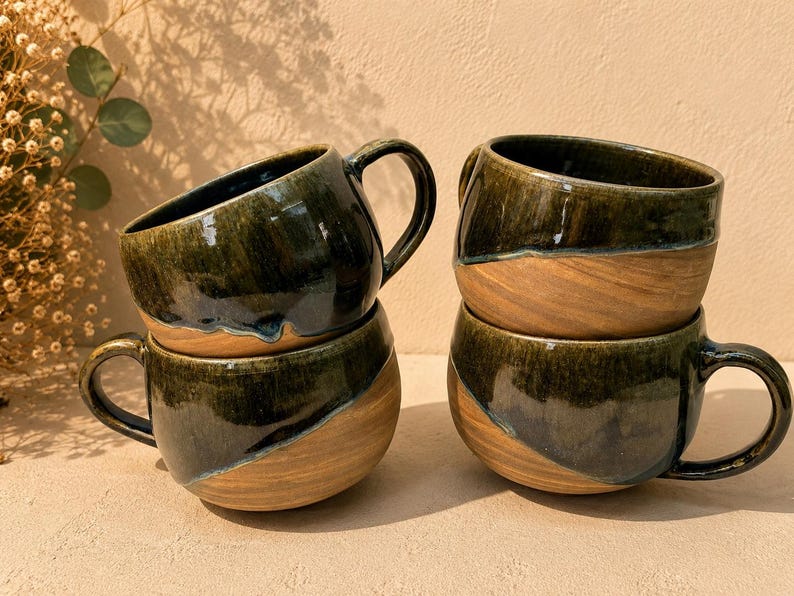 May include: Four ceramic mugs with a rounded shape and a handle on the side. Each mug features a two-tone design with a glossy, dark forest green glaze on the upper portion and a natural, unglazed tan clay finish on the lower portion.