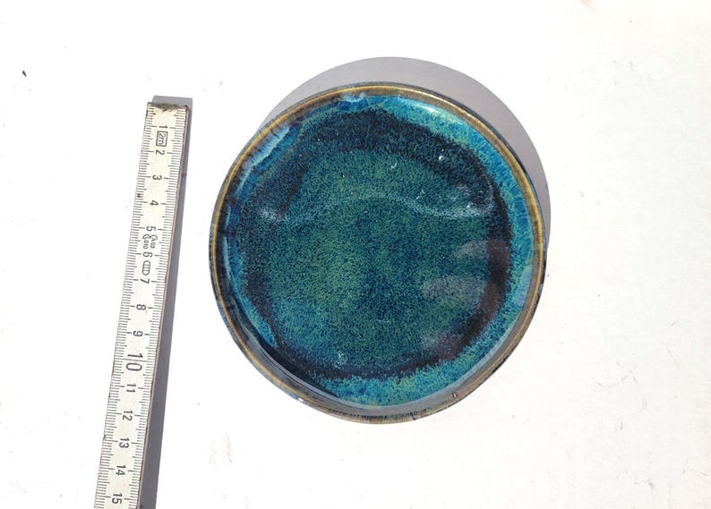 May include: Round ceramic dish with a teal and green glaze. The dish has a dark rim and a textured surface. A ruler is placed next to the dish, showing measurements in inches.