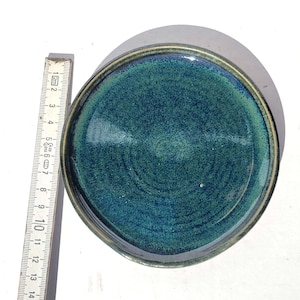 May include: A handmade ceramic plate with a teal and green glaze. The plate has a dark rim and measures approximately 5.5 inches in diameter. The plate's surface has a textured, swirling pattern.