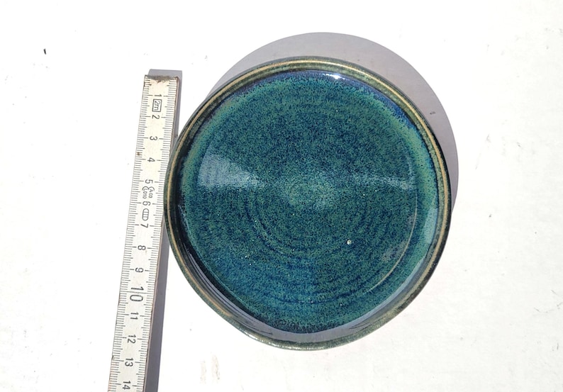 May include: A handmade ceramic plate with a teal and green glaze. The plate has a dark rim and measures approximately 5.5 inches in diameter. The plate's surface has a textured, swirling pattern.