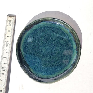 May include: A handmade ceramic plate with a teal and dark green glaze. The plate is round with a slightly irregular edge. A ruler next to the plate shows a measurement of approximately 5 inches in diameter.
