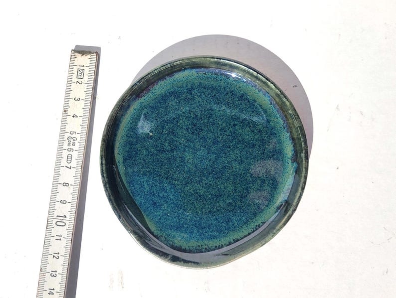 May include: A handmade ceramic plate with a teal and dark green glaze. The plate is round with a slightly irregular edge. A ruler next to the plate shows a measurement of approximately 5 inches in diameter.