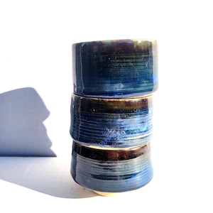 Op de afbeelding: Een stapel van drie handgemaakte keramische kommen. De kommen zijn geglazuurd in blauwe en bruine tinten, met een getextureerd, geribbeld oppervlak. De kommen zijn op elkaar gestapeld.