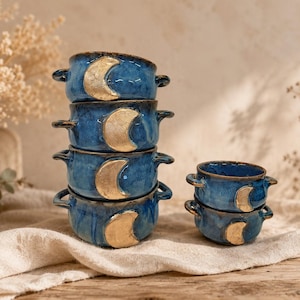 Könnte beinhalten: Ein Stapel aus vier blauen Keramikschalen und ein separater Stapel aus zwei kleineren blauen Keramikschalen. Jede Schale hat zwei seitliche Griffe und ein goldenes Mondsichel-Motiv auf der Vorderseite. Die Glasur ist glänzend und marmoriert.