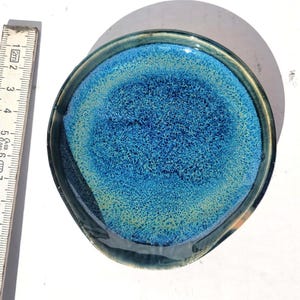 May include: A handmade ceramic spoon rest with a unique blue and green glaze. The spoon rest is round with a small lip and measures approximately 5 inches in diameter. The interior has a textured appearance.