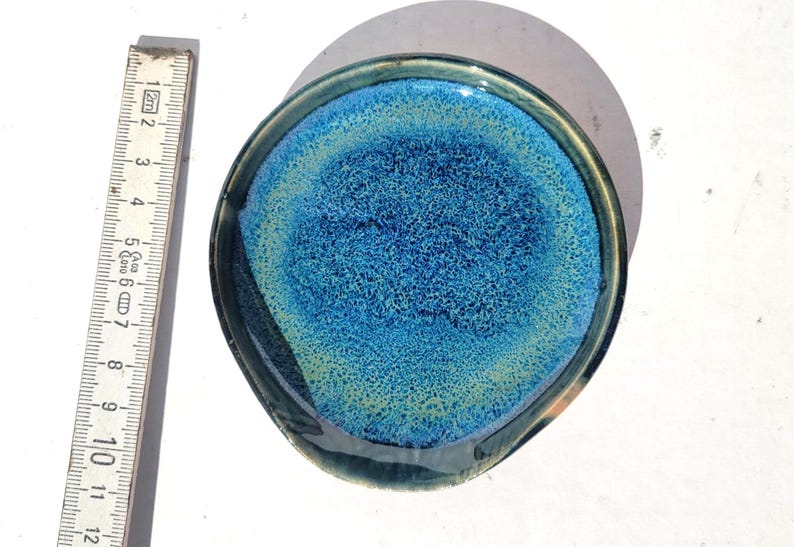 May include: A handmade ceramic spoon rest with a unique blue and green glaze. The spoon rest is round with a small lip and measures approximately 5 inches in diameter. The interior has a textured appearance.