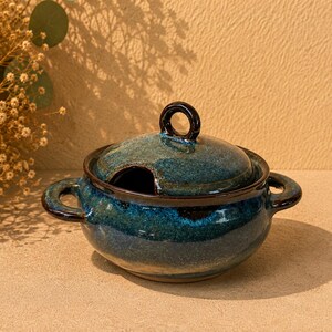 May include: A round ceramic lidded bowl featuring a deep blue and teal mottled glaze. The bowl has two side handles and a matching lid with a circular handle on top. A small notch in the lid allows for a spoon to rest inside the container.