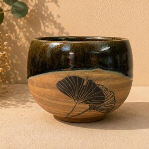 May include: A rounded ceramic bowl featuring a two-tone glaze. The upper portion is a glossy dark brown, while the lower section is a matte tan with a textured surface. A black line drawing of two ginkgo leaves is etched onto the front side.