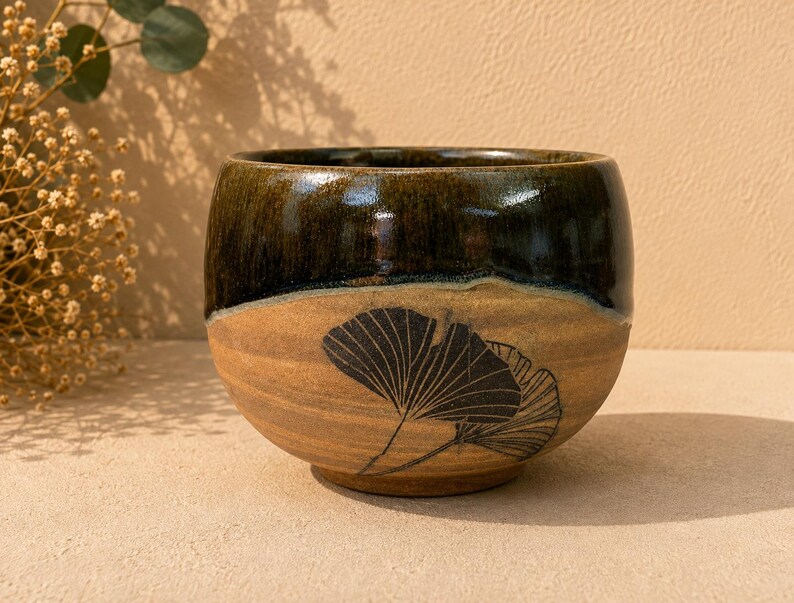May include: A rounded ceramic bowl featuring a two-tone glaze. The upper portion is a glossy dark brown, while the lower section is a matte tan with a textured surface. A black line drawing of two ginkgo leaves is etched onto the front side.