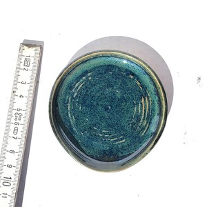 May include: A small, round ceramic dish with a teal interior and a gold-colored rim. The dish has a textured surface and is approximately 4 inches in diameter. The dish is handmade and has a unique, artisanal appearance.