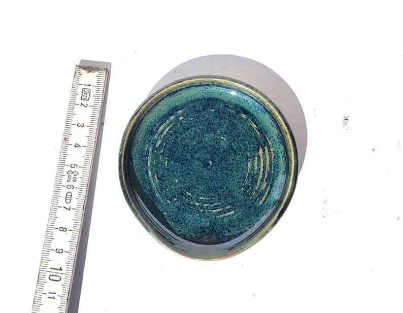 May include: A small, round ceramic dish with a teal interior and a gold-colored rim. The dish has a textured surface and is approximately 4 inches in diameter. The dish is handmade and has a unique, artisanal appearance.