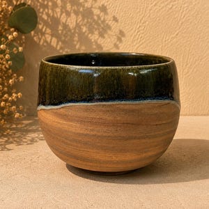 May include: A rounded ceramic bowl featuring a two-tone design. The upper portion is finished with a glossy, dark forest green glaze, while the lower section displays the natural, unglazed tan clay body with visible horizontal striations.
