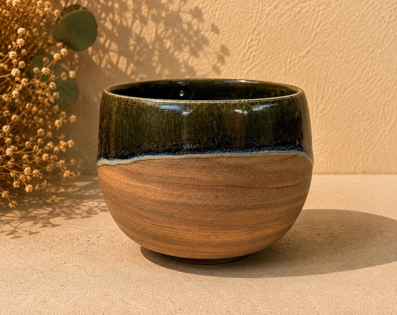 May include: A rounded ceramic bowl featuring a two-tone design. The upper portion is finished with a glossy, dark forest green glaze, while the lower section displays the natural, unglazed tan clay body with visible horizontal striations.
