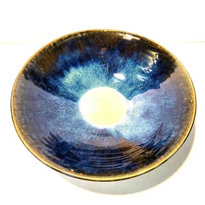 Könnte beinhalten: Eine runde Keramikschale mit glänzendem, schillerndem Finish. Die Schale weist einen Farbverlauf auf, darunter tiefe Blautöne, einen goldbraunen Rand und einen cremefarbenen Mittelbereich. Das Design der Schale deutet auf ein himmlisches oder kosmisches Thema hin.