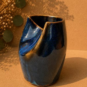 Könnte beinhalten: Eine tiefblaue Keramikvase mit glänzender Oberfläche und einem markanten, gefalteten, asymmetrischen Rand. Das Gefäß zeigt dunkelblaue und kobaltfarbene Töne mit subtilen braunen Akzenten an den Kanten, die eine geschwungene Form bilden.