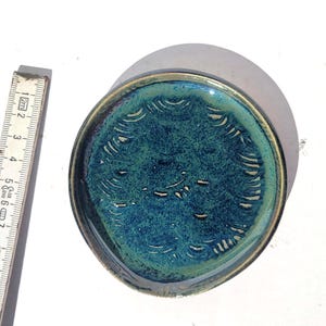 May include: A handmade ceramic dish with a teal and blue glaze. The dish has a circular shape with a diameter of approximately 4 inches. The rim is a darker shade of blue. The interior features a textured pattern.