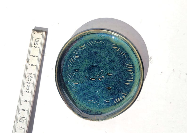 May include: A handmade ceramic dish with a teal and blue glaze. The dish has a circular shape with a diameter of approximately 4 inches. The rim is a darker shade of blue. The interior features a textured pattern.