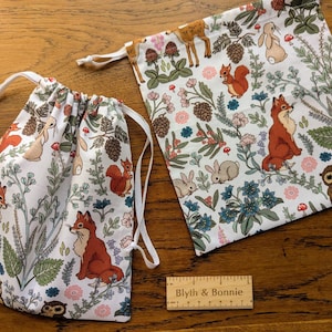 以下が含まれることがあります： 森の動物柄の白い巾着袋が2つ。プリントには、キツネ、リス、ウサギ、フクロウが花や葉の間に入っています。袋は布製で、白い紐が付いています。袋は木製の表面にあります。