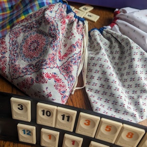 May include: A selection of fabric drawstring bags with diverse patterns, including paisley and floral designs. The bags are in shades of white, red, and blue. Rummikub game tiles are visible in the foreground.