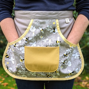 Op de afbeelding: Een grijze schort met een bijen- en bloemenpatroon, met een gele zak en afwerking. De schort heeft twee grote zakken en een tailleband. Het ontwerp bevat zwart-gele bijen en witte bloemen.