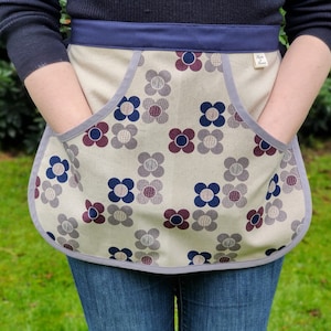 Puede incluir: Delantal beige con un estampado floral en tonos azules, burdeos y grises. El delantal tiene dos bolsillos grandes y una cinturilla azul marino. Los bordes están ribeteados con tela gris. Una pequeña etiqueta está adherida al delantal.