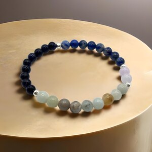 May include: A beaded bracelet featuring a spectrum of colors. The bracelet is composed of various round beads in shades of dark blue, light blue, green, gray, and lavender. Silver-colored spacers are interspersed between the beads. The bracelet is displayed on a gold-colored surface.