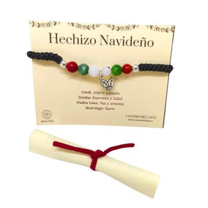 Op de afbeelding: Een kerst-geluksarmband met rode, groene en witte kralen, een zilveren bedel met een hulsttak en een zwart koord. De armband wordt gepresenteerd op een kaart met de tekst "Hechizo Navideño" en informatie over de stenen en hun betekenis.