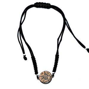 Op de afbeelding: Zwart gevlochten koordarmband met een ronde hanger met een vijfpuntige ster met een symbool erin. De hanger is bruin met een zilveren rand.