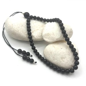 Op de afbeelding: Een zwarte kralenketting met een schuifknoopsluiting. De ketting bestaat uit kleine, ronde, gepolijste zwarte kralen geregen aan een zwart koord. De ketting wordt gepresenteerd op witte, gladde stenen.