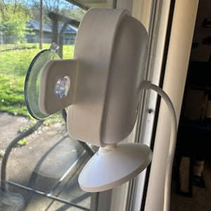 Puede incluir: Una cámara de seguridad blanca con un soporte de ventosa fijado a una ventana. La cámara tiene un cable blanco.