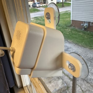 May include: White security camera mounted on a window with suction cups. The camera has a speaker grille and a cord.