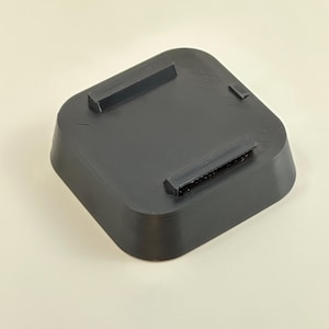 May include: A black, square-shaped 3D-printed object with rounded corners and raised rectangular supports on the top surface. The object is made of a matte black material and sits on a neutral background.