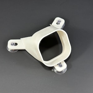 Puede incluir: Un dispositivo de plástico blanco con una abertura cuadrada y tres ventosas. El dispositivo está diseñado para adherirse a una superficie lisa. El objeto está hecho de plástico liso y mate.