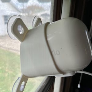 Pode incluir: Uma câmera de segurança branca com um suporte de ventosa preso a uma janela. A câmera está conectada a um cabo de alimentação e tem a palavra "arlo" impressa no lado.