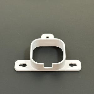 May include: A white, 3D-printed bracket with a rectangular opening and a small loop at the top. The bracket has a flat base with two circular holes. The object is designed to hold something, possibly a small electronic device.
