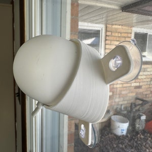 May include: White 3D printed bird feeder with a suction cup mount attached to a window. The feeder has a small spout for birds to eat from.