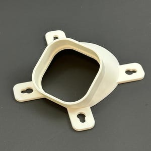 Puede incluir: Un objeto de plástico blanco de forma cuadrada, con una abertura central y cuatro pestañas salientes, cada una con un orificio en forma de ojo de cerradura. Probablemente sea un soporte de montaje.