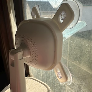 May include: White security camera with a round base and four suction cups attached to a window. The camera has a speaker grille and a swiveling head.