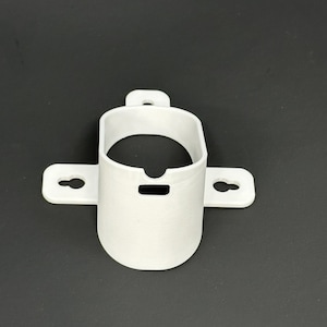 May include: A white, 3D-printed bracket with a cylindrical body and two horizontal wings, each with a keyhole-shaped opening. The bracket has a small, rectangular opening at the top and a small loop at the top for hanging. The object is set against a dark background.