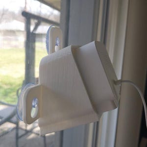 Może przedstawiać: Białe, drukowane w 3D urządzenie z prostokątną podstawą i skośną górą, przeznaczone do trzymania białego urządzenia elektronicznego. Urządzenie jest przymocowane do okna za pomocą dwóch przyssawek. Biały kabel wychodzi z urządzenia.